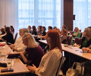 Na fotografii sú účastníci a účastníčky konferencie. Sedia na stoličkách a pozerajú dopredu či do papierov na stole pred sebou
