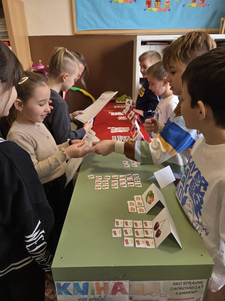 Deti stoja okolo stola v školskej triede, pozerajú do papierov a na seba a plnia aktivitu.