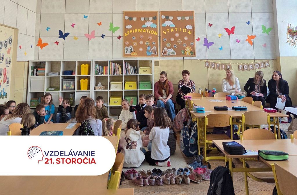 Na fotografii je školská triede, kde sedia vzadu na stoličkách učitelia a učiteľky z iných škôl počas EDUKAfestu 2026 a pozerajú sa na deti a pani učiteľku, ktorá vedie otvorenú hodinu a spolu s deťmi sedí na koberci.